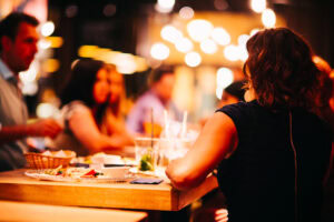A woman with short hair sits at a wooden table in a dimly lit restaurant, facing a group of people. Food, drinks, and plates are on the table, with warm string lights blurred in the background.