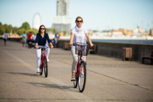 Two women riding bicycles along a wide, sunny promenade near the water, with buildings, a ferris wheel, and trees visible in the background.