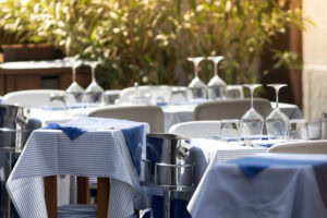 Several tables at an outdoor restaurant are set with blue and white striped tablecloths, upturned wine glasses, and cutlery, with sunlight filtering through leafy plants in the background.
