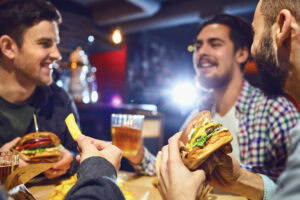 Three men are laughing and talking at a restaurant table, holding burgers and beer, while another hand is holding a french fry in the foreground. The setting appears relaxed and casual.