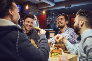 Four young men sit together at a bar table, laughing and talking while enjoying burgers and drinks. The atmosphere is casual and friendly, with warm lighting and bar decor in the background.