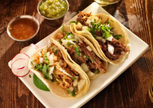 Three tacos filled with shredded meat, chopped onions, and cilantro on a white plate, served with sliced radishes, lime, and two cups of salsa on a wooden table.