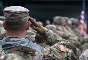 A group of soldiers in camouflage uniforms, one in the foreground saluting with a gloved hand. An American flag patch is visible on a sleeve, and a U.S. flag appears blurred in the background.