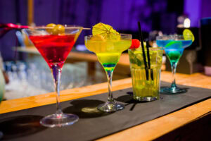 Four colorful cocktails in various glasses are lined up on a bar; each drink is garnished differently, including olives, a paper umbrella, fruit, and a lime, set against a blurred background with purple lighting.