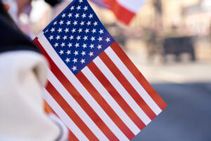 A close-up of a small American flag being held outdoors, with the stars and stripes clearly visible. The background is blurred, suggesting a public event or parade.
