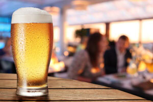 A cold, frothy pint of beer sits on a wooden table, covered in condensation, with a blurred background of people talking and dining in a warmly lit restaurant or bar.