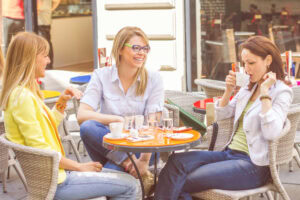 Three women sit at a café table outdoors, smiling and chatting. Coffee cups, glasses of water, and smartphones are on the table. They appear relaxed and engaged in conversation on a sunny day.
