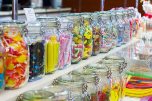 Rows of glass jars filled with colorful assorted candies and sweets are displayed on shelves in a candy store. Some jars are open, showing gummy bears, licorice, jelly beans, and sour belts.