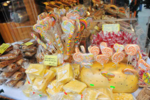 A market stall displays colorful swirled lollipops, large blocks of cheese, and packaged pastries. Signs with handwritten labels are visible among the various treats.