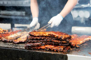 A person wearing gloves grills several racks of barbecue ribs, with a smoky atmosphere and a large stack of cooked ribs in the foreground.