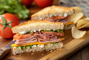 A ham and cheese sandwich with lettuce, tomato, and mayonnaise on sliced bread, cut in half and stacked, sits on a wooden board beside potato chips, fresh tomatoes, and lettuce in the background.