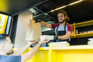 A smiling food truck worker in an apron hands a paper bag and a food container to a customer over a yellow counter. The worker is wearing black gloves and the interior of the truck is also yellow.