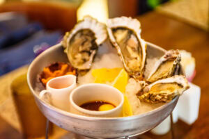 A metal dish filled with ice holds four fresh oysters on the half shell, a lemon wedge, and two small cups with sauces, set on a wooden table in a restaurant setting.