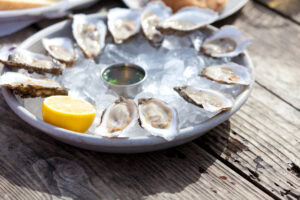 A plate of raw oysters served on a bed of ice with a lemon half and a small container of dipping sauce, set on a rustic wooden table.