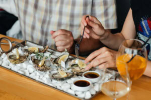 Two people are sitting at a table, sharing a tray of oysters on ice with lemon wedges and sauces. One person is using a fork, and two drinks are visible in the foreground.