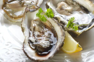 Close-up of fresh oysters on the half shell, garnished with sprigs of parsley and served with a lemon wedge on a white plate. The oysters are glistening with their natural juices.