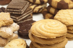 An assortment of cookies and biscuits, including round butter cookies, chocolate wafers, and checkerboard-patterned pieces, stacked and arranged closely together on a white surface.