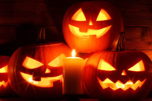 Four glowing jack-o-lanterns with carved faces sit behind a lit candle, casting an orange light against a dark wooden background, creating a spooky Halloween atmosphere.