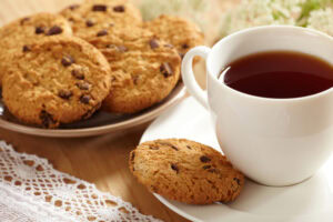 A white cup filled with tea sits on a saucer beside a chocolate chip cookie. In the background, a plate holds several more cookies, all on a wooden table with a lace tablecloth and white flowers nearby.