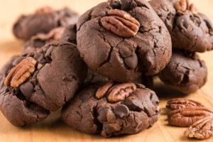 A close-up of several chocolate cookies topped with pecan halves and chocolate chips, stacked on a wooden surface.