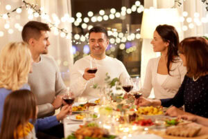 A group of six people sit around a festive dinner table, smiling and holding glasses of wine, with plates of food and decorative lights creating a warm, joyful atmosphere.