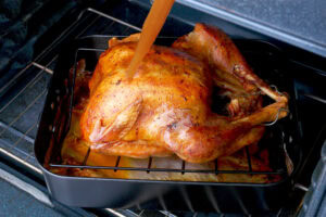 A whole roasted turkey sits in a pan on an oven rack while a hand pours broth or juices over it to keep it moist. The turkey’s skin is golden brown and crispy.