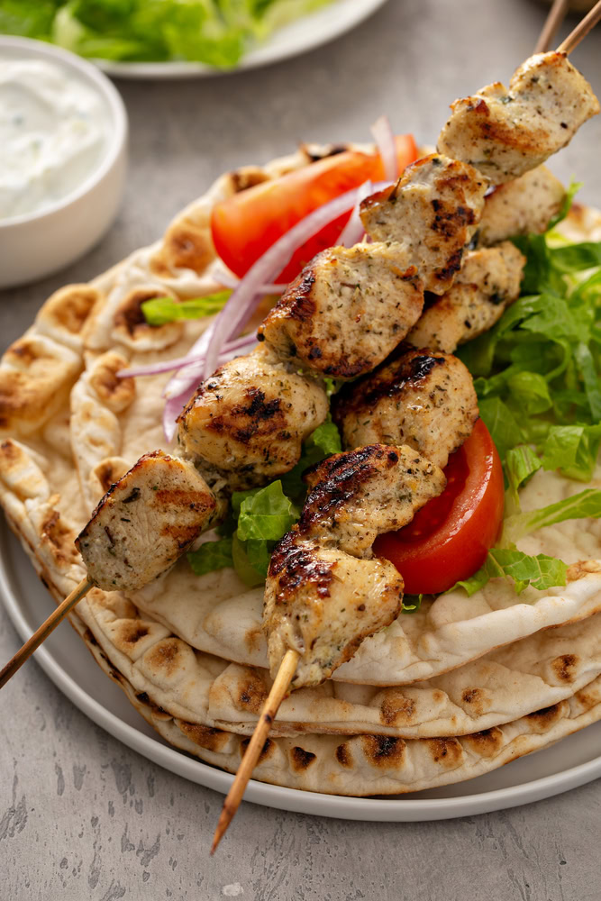 Two grilled chicken skewers are placed on top of warm pita bread, garnished with fresh lettuce, tomato slices, and red onion. A bowl of white sauce and a plate with more greens are in the background.