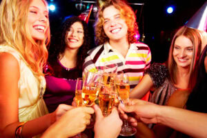 A group of young adults smiling and toasting with glasses of champagne at a lively party, wearing party hats and festive outfits, with colorful lights in the background.