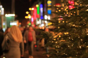 A decorated Christmas tree with glowing lights is in focus on the right, while a busy city street with blurred people and colorful illuminated signs is seen in the background at night.