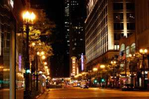 Nighttime city street scene with bright streetlights, illuminated buildings, American flags, and a vertical CHICAGO theater sign glowing in the background amid downtown traffic.