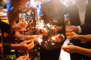 A group of people in festive attire hold sparklers and glasses of champagne, celebrating together in a lively, warmly lit setting.
