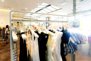 A rack of various women’s clothing hangs in a bright, modern boutique with wooden floors and large windows. A person walks in the background. The shop is well-lit and spacious.