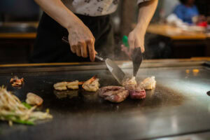 A chef cooks shrimp, steak, and vegetables on a hot teppanyaki grill, using two metal spatulas. Steam rises from the sizzling food, and diners are blurred in the background.