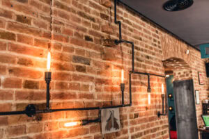 Exposed brick wall with industrial-style lighting made from metal pipes and vintage bulbs, arranged in a geometric pattern along the wall.