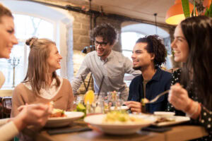 A group of young adults sit at a restaurant table, smiling and talking with a waiter standing nearby. Plates of food and drinks are on the table, and the atmosphere is warm and friendly.