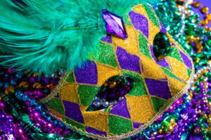 A glittery Mardi Gras mask with green feathers and a purple jewel rests on a pile of colorful beads in purple, green, and gold. The mask features a diamond pattern in purple, green, and yellow.