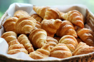 A basket filled with golden brown, flaky croissants, arranged closely together on white parchment paper. The croissants have a shiny, crisp outer layer and are in a woven basket.