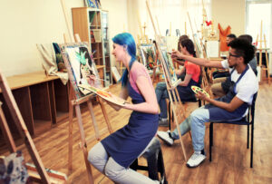 A group of people wearing aprons sits at easels painting in a bright art classroom. The focus is on a young woman with blue hair painting a still life, surrounded by other students working on their own canvases.