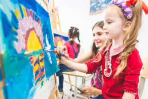 A young girl in a red shirt paints a colorful sunset on an easel, guided by a smiling woman. Another child paints in the background, also focused on her artwork. The setting appears to be an art classroom.