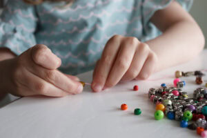 A child’s hands string colorful beads on a table. Several loose beads are scattered on the white surface. The child is wearing a blue top with a wavy pattern.