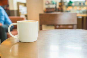 A white coffee mug sits on a round wooden table in a cafe, with a blurred background showing an empty chair and a person partially visible on the left.