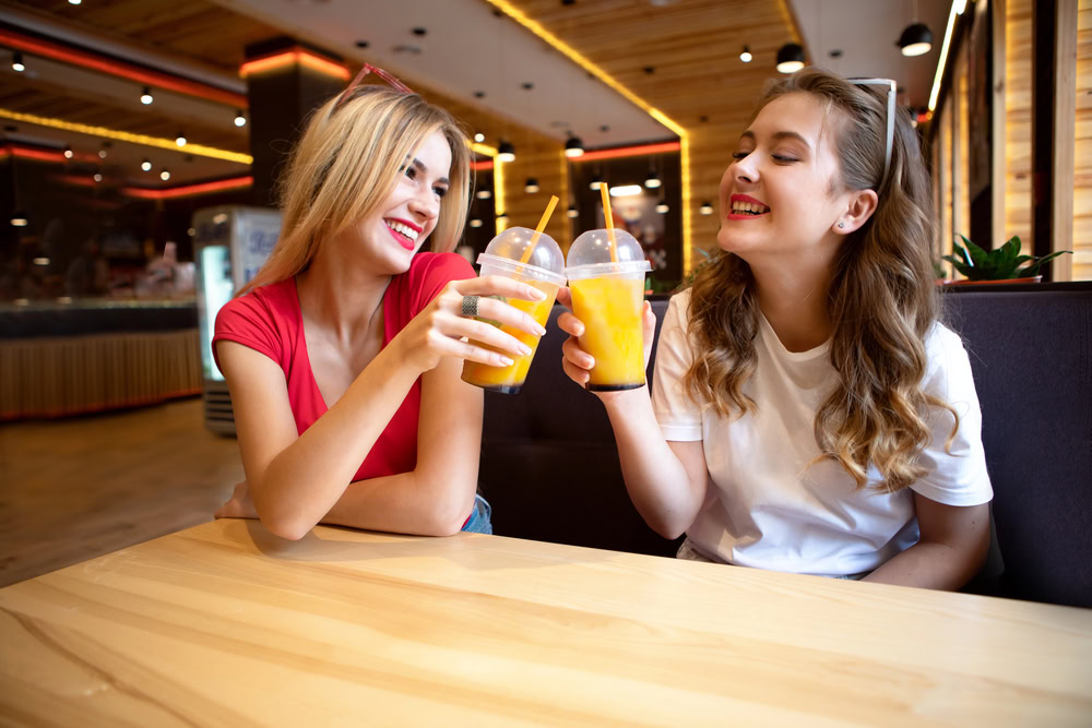 Two young women smiling and sitting at a wooden table in a modern cafe, toasting with plastic cups of orange juice.