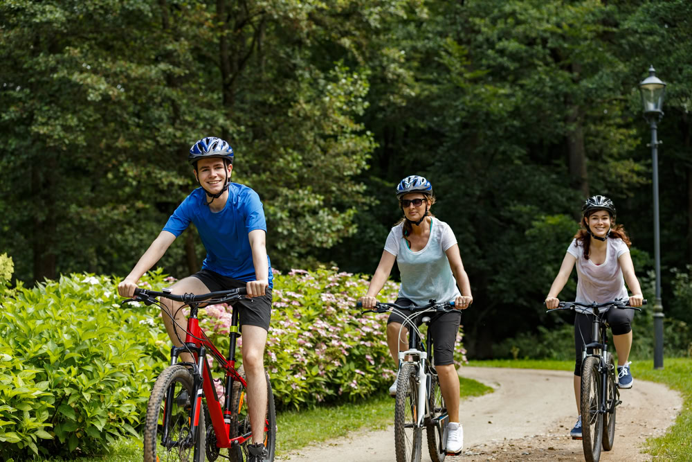 Three people wearing helmets ride bicycles along a curved path through a green park, surrounded by trees and bushes, on a sunny day. They are smiling and appear to be enjoying the outdoors.