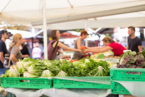 A farmers market scene with fresh leafy greens in green crates in the foreground, and several people shopping and exchanging goods under white tents in the background.