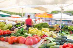 Fresh vegetables including tomatoes, bell peppers, zucchini, and cucumbers are displayed on a market stall under large umbrellas, with people blurred in the background.