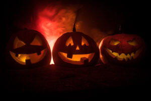 Three carved jack-o’-lanterns with glowing faces sit in a row on dark ground. A red, smoky light glows behind them, creating a spooky Halloween atmosphere.