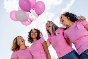Four women wearing pink shirts and pink ribbon pins smile and laugh together outdoors, holding pink and white balloons, with a bright sky in the background.