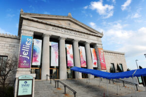 Exterior of the John G. Shedd Aquarium with large columns, colorful banners advertising a Jellies exhibit, a sign near the entrance, and a blue awning over the steps under a partly cloudy sky.