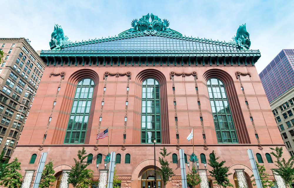 The image shows the exterior of the Harold Washington Library Center in Chicago, featuring large arched windows, red stone walls, and a green ornamental roof with sculpted owls and foliage. Two flags are flying in front.