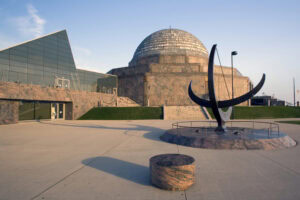 A large stone sundial stands in front of a modern glass building and a domed observatory, under a clear blue sky. The setting appears open and spacious with concrete walkways.